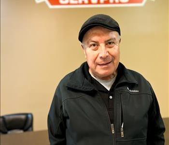 male employee with black hat and jacket in front of brown wall with company logo